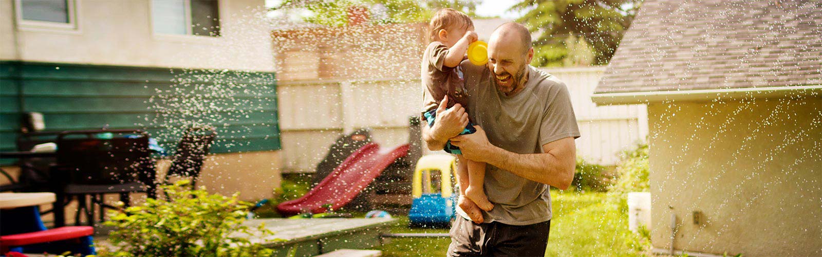 Father carrying son through water sprinkler backyard building